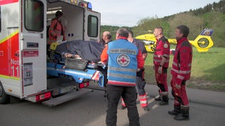 Eine Gruppe von Rettungskräften, darunter auch Personal der Heli-Retter, steht vor einem Krankentransport. Eine Trage wird eingeladen. Im Hintergrund steht der gelbe Helikopter "Christoph 22".