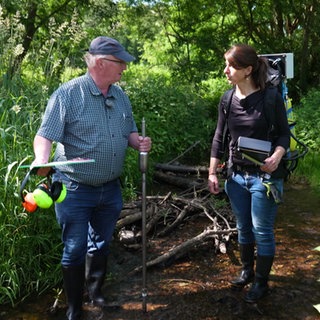 Gewässerökologe Dr. Hans-Jürgen Hahn und Dr. Heide Stein stehen im Guldenbach mit einem Messgerät.