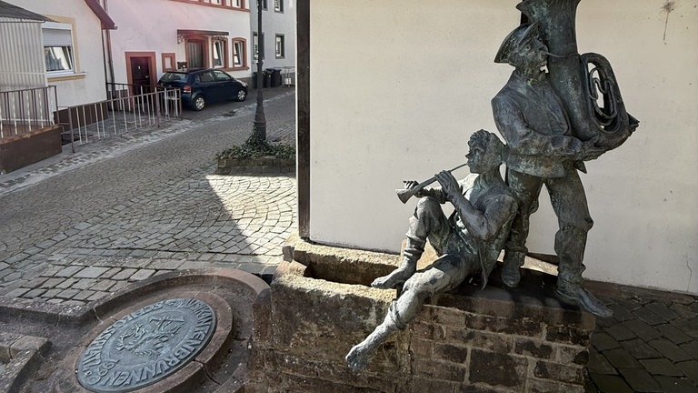 Ein Brunnen in Wolfstein mit zwei menschlichen Figueren, die Blasinstrumente spielen. Er soll an die Tradition der Wandermusikanten erinnern.