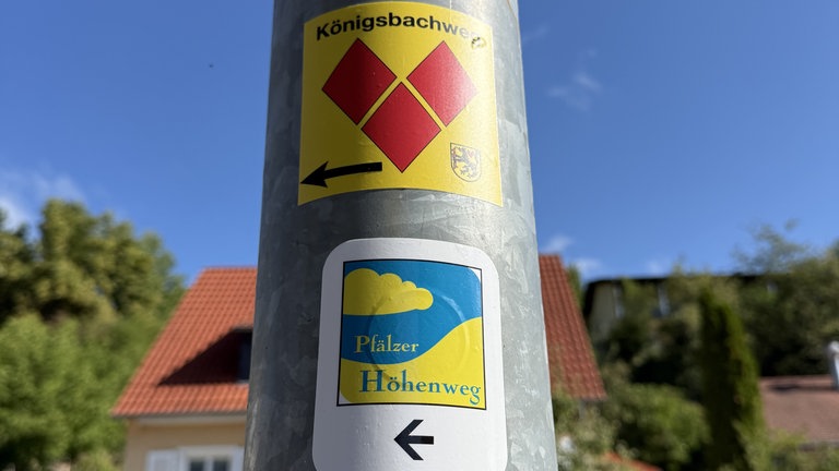 Zwei Aufkleber auf einem Laternenpfahl, die als Wegmarkierungen für Wanderwege dienen, darunter auch der Königsbachweg. Er wird dargestellt mit drei roten Rautensteinen auf gelbem Grund.