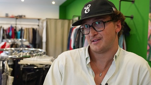 Jason Bonk sitzt in seinem Second-Hand-Laden in Koblenz vor Kleiderstangen.