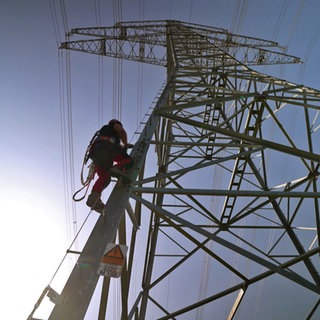 Bauarbeiter mit Helm beobachtet einen Kollegen beim Besteigen eines Strommasts