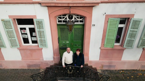 Ein älterer Mann und eine ältere Frau stehen auf der Treppe vor dem Eingang eines alten, imposanten Hauses.