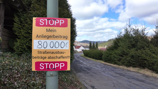 Auf einem Schild steht: "STOPP - Mein Anliegerbetrag 80.000€ - Straßenausbau-beiträge abschaffen!"