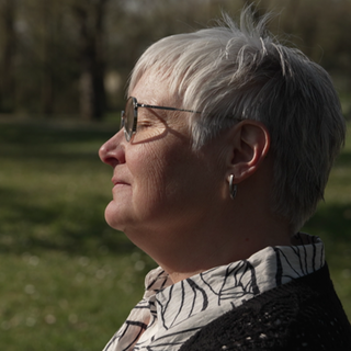 Anke Domay sitzt mit geschlossenen Augen auf einer Parkbank, die Sonne scheint auf ihre ruhigen Gesichtszüge.