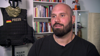 Mann sitzt vor einem Regal und einer Schutzweste.