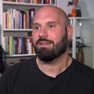 Mann sitzt vor einem Regal und einer Schutzweste.