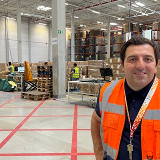 Logistiker Kreshnik Himaj steht lächelnd in einer Lagerhalle vor gestapelten Paketen.