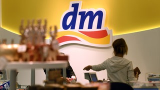 Junge Frau - man sieht sie von hinten - die gerade in einem dm-Markt vor dm-Logo ihre Ware bezahlt. Wie gut ist dm? Marktcheck testet die Eigenmarke Balea und guckt auf die Preise im Vergleich zu Rossmann und Müller.