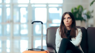 Eine Frau sitzt beleidigt mit ihrem Koffer am Flughafen. Gepäck verloren auf der Reise oder schlechtes Hotel: Ihre Rechte im Urlaub bei Reisemängeln und welche Entschädigung Ihnen zusteht.