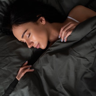 Junge Frau mit dunklen Haaren und zufriedenem Lächeln schläft unter dunkler Bettdecke. Helfen Gewichtsdecken bei Schlafproblemen?