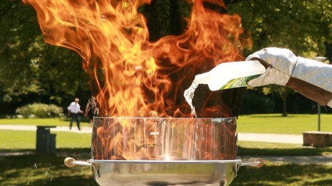 Feuerwehrmann in Schutzanzug schüttet Spiritus auf brennenden Grill mit Stichflamme. Erste Hilfe bei Verbrennungen: Soll man Brandwunden kühlen?