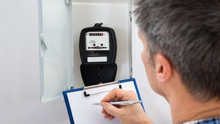 Ein Mann hält ein Klemmbrett und einen Stift in der Hand, als wolle er etwas notieren und schaut dabei auf einen Stromzähler.