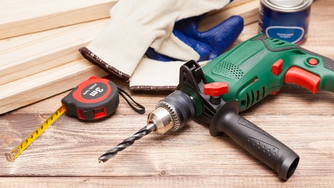 Akku-Bohrmaschine, Zollstock, Arbeitshandschuhe, Leimdose und Holzbretter liegen auf einem Tisch