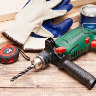 Akku-Bohrmaschine, Zollstock, Arbeitshandschuhe, Leimdose und Holzbretter liegen auf einem Tisch