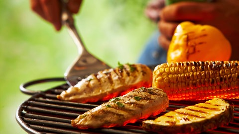 Auf einem Grillrost liegen ein Maiskolben, zwei Steks und eine Aubergine. Eine Hand hat einen Grillwender in der Hand und hebt damit ein Steak ab.