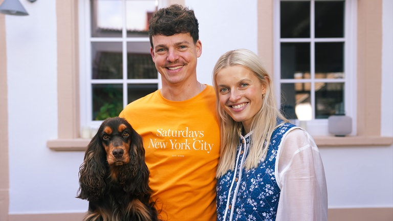 Peter mit Hund auf dem Arm und Franziska stehen lächelnd vor ihrem kleinen Landhäuschen.
