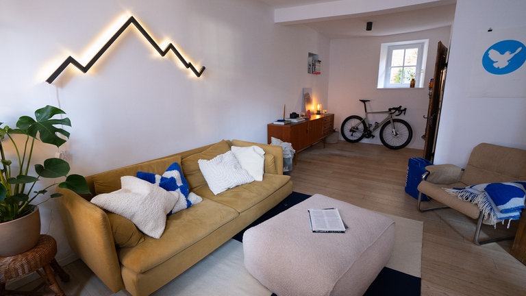 Ein gemütliches Wohnzimmer mit einer senfgelben Ikea-Couch, Kissen und einer modernen, auffälligen Wand-Lampe in Form eines Bergmassivs
