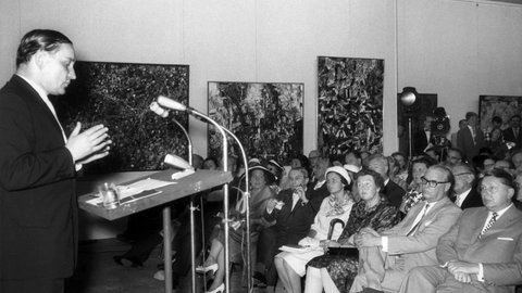 Werner Haftmann spricht während der Eröffnung der documenta 2 im Jahr 1959 in Kassel. Im Publikum vorn mit Brille der hessische Ministerpräsident Georg August Zinn mit Gattin und rechts daneben der Initiator der documenta, Arnold Bode