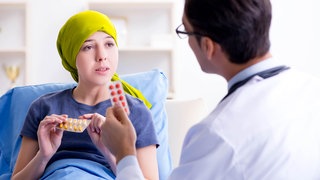 Frau mit Glatze unter ihrem gelben Kopftuch redet mit Arzt über ihre Chemotherapie: Eine Chemotherapie hat oft Haarausfall als Nebenwirkung